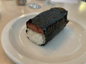 On a white plate is a white rice shaped as a rectangle. On top of the rice is a rectangular shaped, homemade, spam with soy sauce. The stacked rice and spam are wrapped in seaweed.