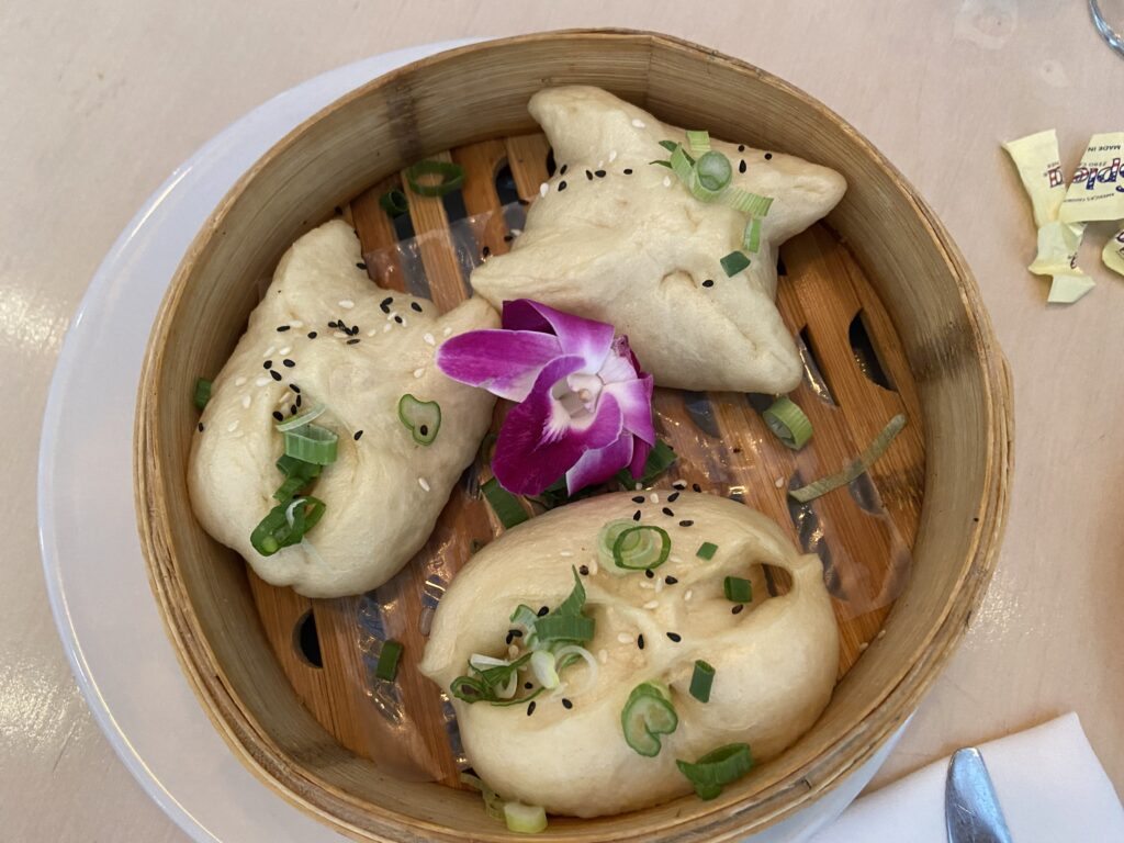 Three steamed Chinese-style buns with black and white sesame seeds and sliced green onion on top and a dark pink flower in the middle in a bamboo steam basket. Inside the steamed buns are char siu pork, onions and cilantro.