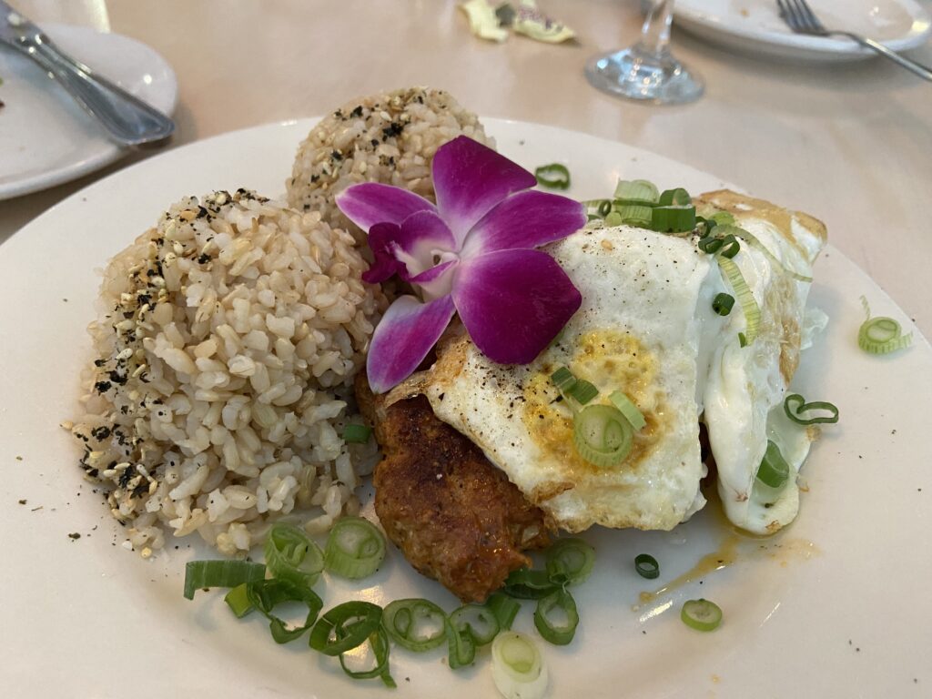 Two portuguese sausage patties with two over easy eggs and two mounds of brown rice with black and white sesames seeds and green onion on top. Garnished with a purple flower. 
