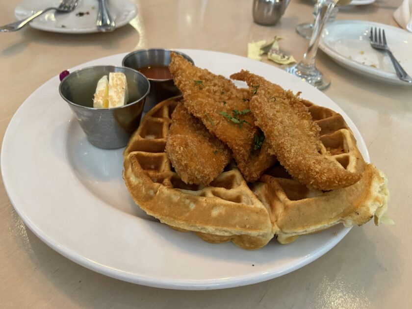Three golden-brown breaded chicken strips on top of a macadamia nut waffle with a red sriracha mango syrup and butter on the side.