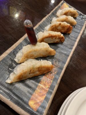Six pan fried potstickers on a rectangle plate. Injected on the second from bottom potsticker is a plastic syringe filled with soy sauce.