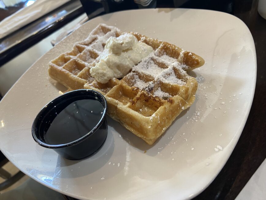 A sweet waffle with powdered sugar and creme on top. And there's a small side of syrup.