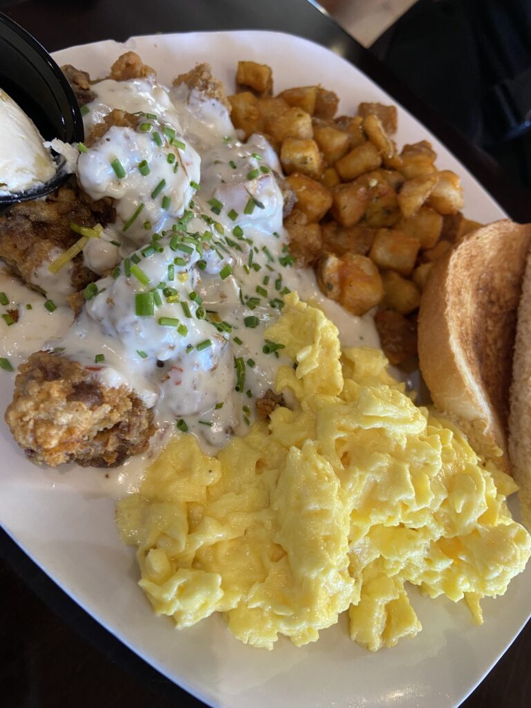 Chicken Fried Steak with a white sauce and chopped chives on top. The sides are scrambled eggs, wheat toast (a side of butter), and roasted potatoes.
