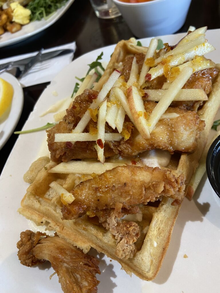 A sweet waffles with butter with fried chicken and aa sweet apple slaw on top. On the side is a bed of arugula and a small cup of syrup.