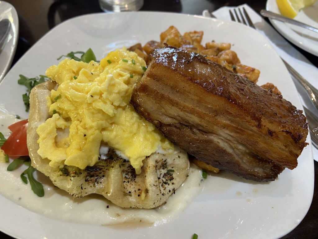 A savory waffle above a bed of arugula and tomato salad. On top are scrambled eggs with chives , and pork belly with a side of roasted potatoes.