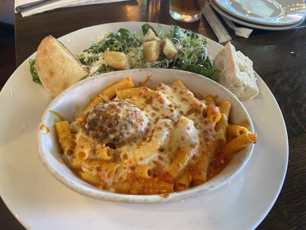 Pasta and Meatball Bake. Ziti pasta baked with marinara sauce  and cheese with a meatball. On the side is two pieces of bread and Caesar salad. 