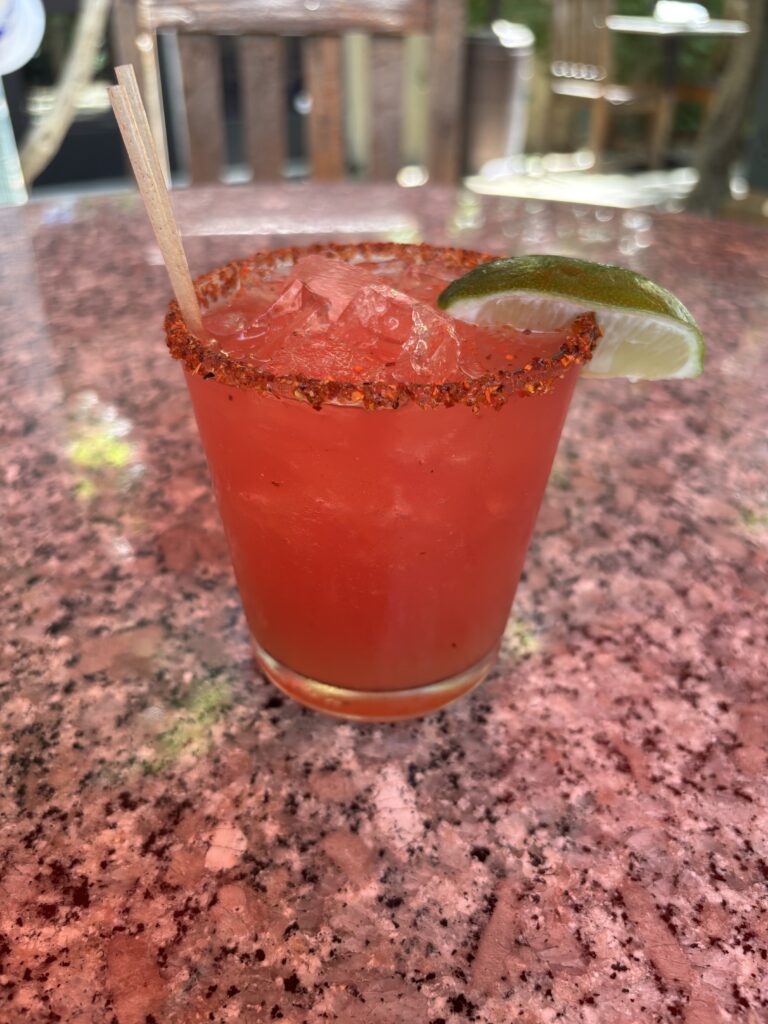 A non-alcoholic drink with stirringd blood orange bitters, agave, blood orange juice, lime juice, with a tajin rim and a lime wedge.