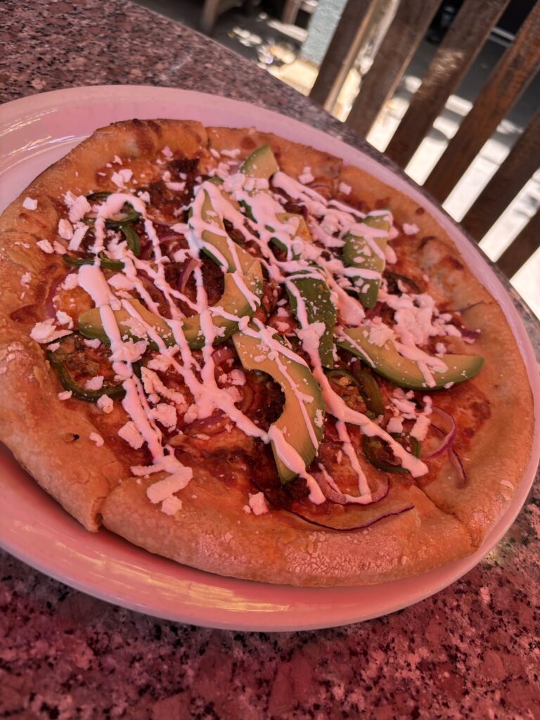 a pizza with chorizo, mozzarella, smoky ancho-pasilla chile sauce, jalapeños, red onions, and cilantro topped with queso fresco, avocado, cream fraiche.