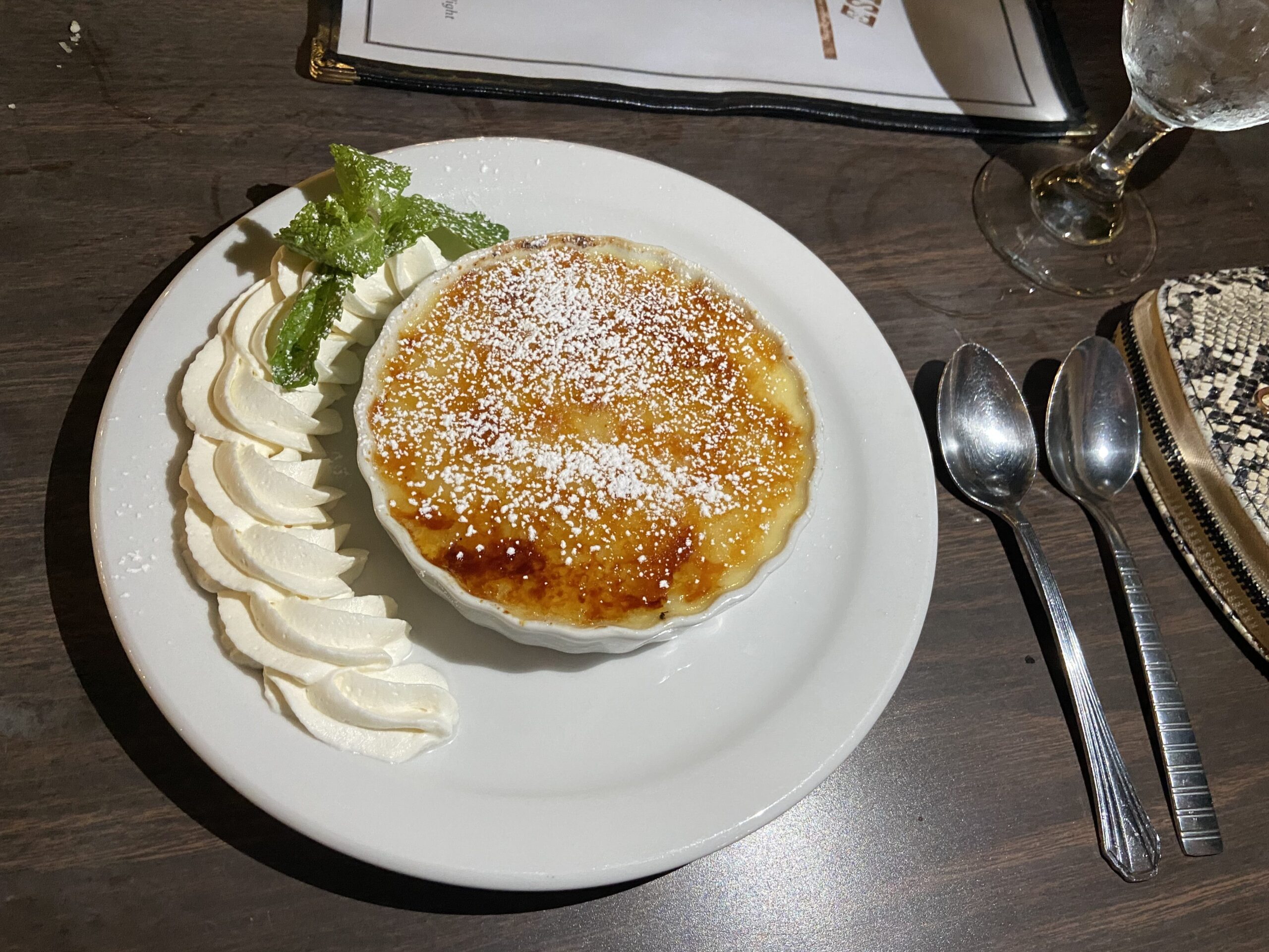In a small ramekin is crème Brûlée. On top of the crème brûlée is powdered sugar. To the right is beautiful crescent line of whipped crème with a sprig of mint.